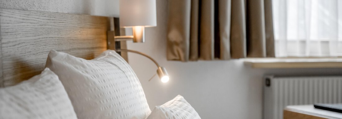 A cozy bedroom with a wooden bed frame and several pillows. Soft curtains and a modern lamp create an inviting atmosphere. - Apart Hotel Garni Wieser - Sölden
