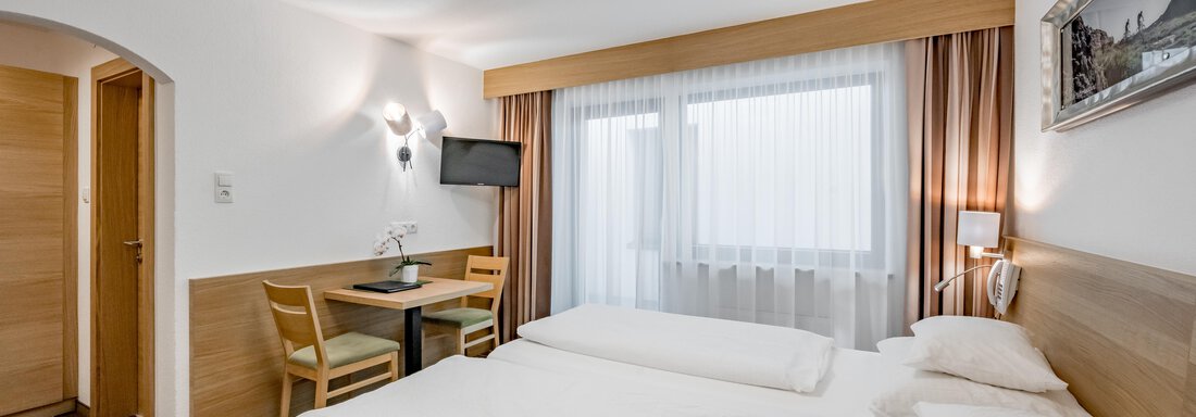 A modern hotel room with two beds, a table, and a television. A lot of daylight comes in through the large windows. - Apart Hotel Garni Wieser - Sölden