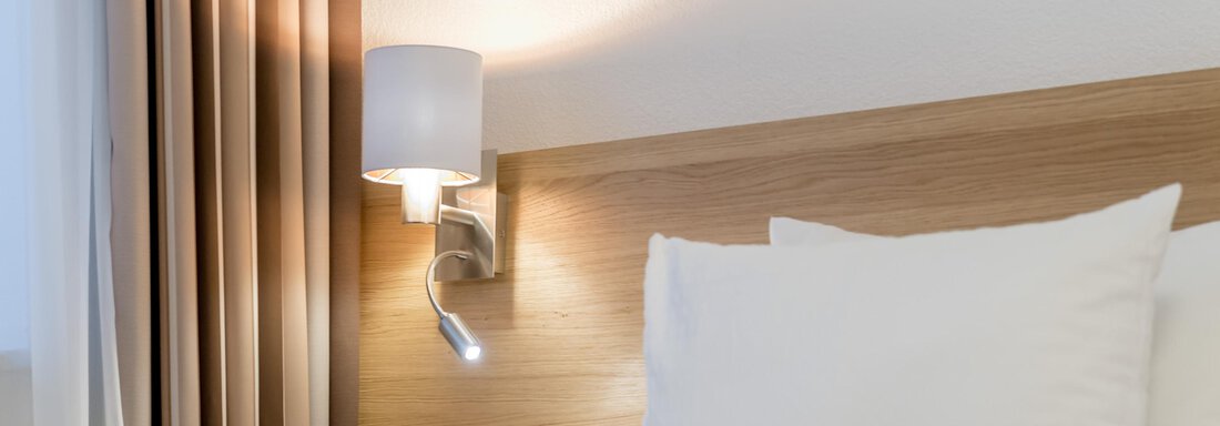 A cozy bedroom with a wooden bed frame and white pillows. A modern lamp is next to the curtains on the wall. - Apart Hotel Garni Wieser - Sölden