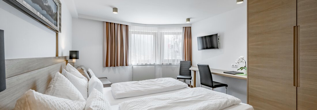 A modern hotel room with two single beds, white bedspreads, and a desk. The room is bright and features large windows as well as a television. - Apart Hotel Garni Wieser - Sölden