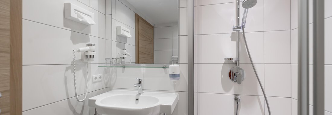 A modern bathroom with white tiles and a shower. There is a sink and a mirror. - Apart Hotel Garni Wieser - Sölden
