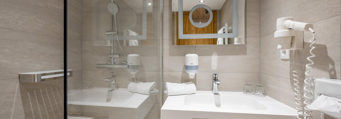 A modern bathroom with a shower, a sink, and a large mirror. The walls are in light tones and the furnishings are functional. - Apart Hotel Garni Wieser - Sölden