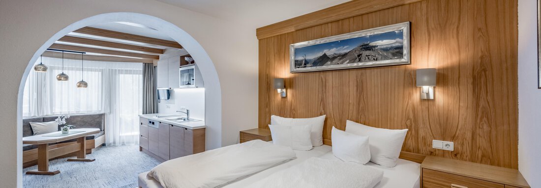 A modern hotel room with a large double bed and warm wooden details. In the background, a living area with a table and a kitchen is visible. - Apart Hotel Garni Wieser - Sölden