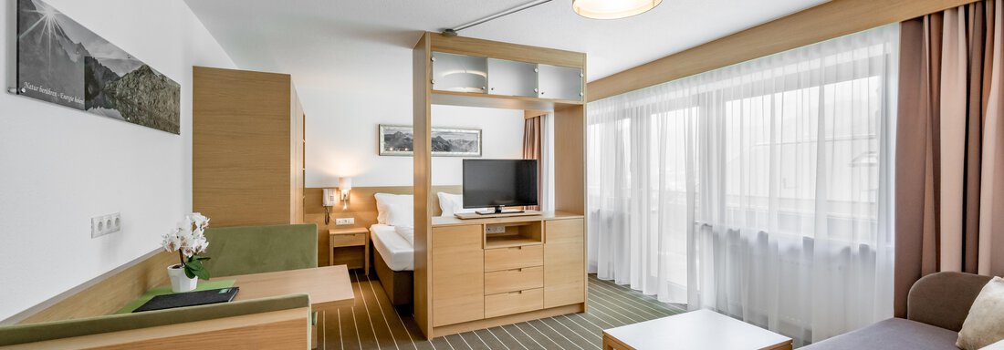 A modern hotel room with bright wood interior and large windows. It features a desk, a television, and a cozy seating area. - Apart Hotel Garni Wieser - Sölden