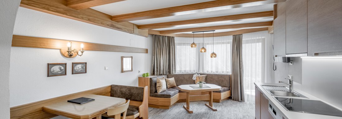A modern living room with wooden ceiling beams and large windows. There is a dining area with a table and chairs, as well as a cozy seating corner. - Apart Hotel Garni Wieser - Sölden