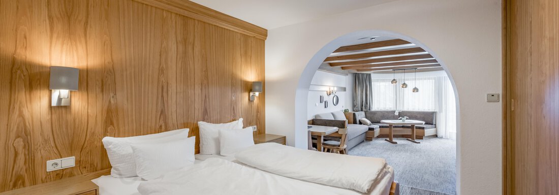 A modern hotel room with wooden paneling, two single beds, and soft lighting elements. In the background, a comfortable living area with large windows can be seen. - Apart Hotel Garni Wieser - Sölden