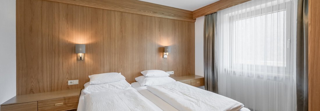 A modern bedroom with two single beds and wooden decor. The room has large windows with bright curtains and natural lighting. - Apart Hotel Garni Wieser - Sölden