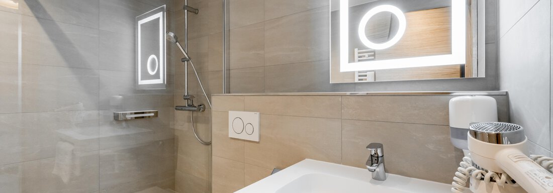 A modern bathroom with bright tiles and a clear glass shower. The sink features an elegant mirror with integrated lighting. - Apart Hotel Garni Wieser - Sölden