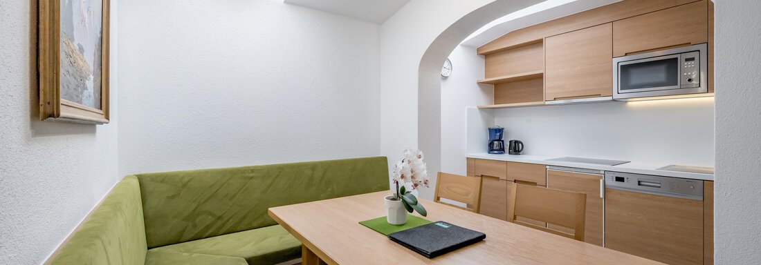 A modern kitchen with light walls and a large window. A green sofa around a wooden table creates an inviting atmosphere. - Apart Hotel Garni Wieser - Sölden