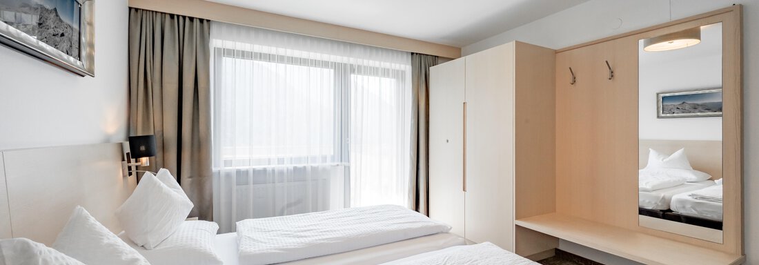 A modern hotel room with two single beds and a large window. The room is bright and cheerfully decorated, with a mirror and a luggage rack. - Apart Hotel Garni Wieser - Sölden