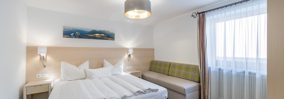 A modern hotel room with a large bed and a sofa. Large windows provide bright daylight. - Apart Hotel Garni Wieser - Sölden