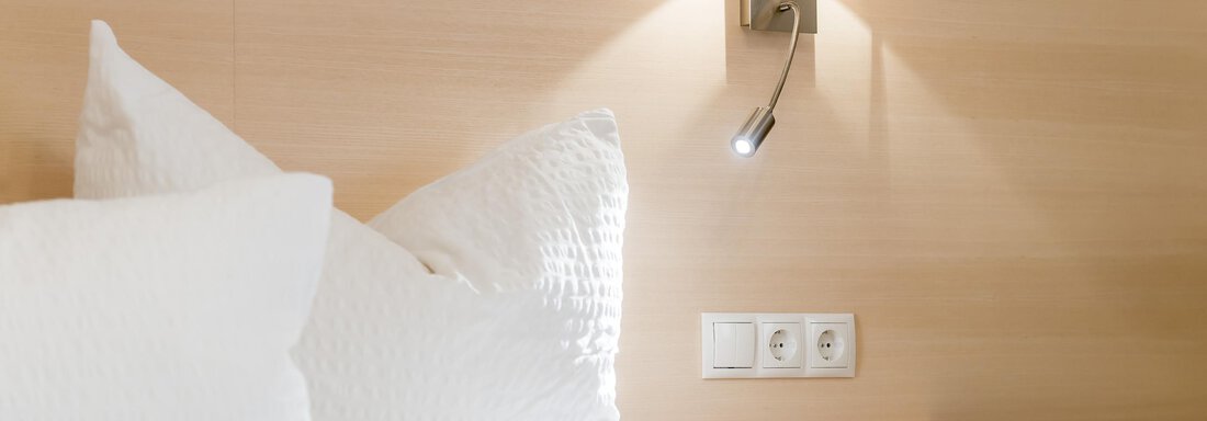 A cozy bed with white pillows and a modern bedside table. On the wall, there is a stylish light source. - Apart Hotel Garni Wieser - Sölden
