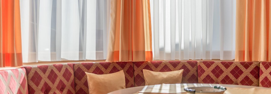 A cozy dining area with a round wooden tabletop and a padded bench. The orange curtains and the lamp create a warm atmosphere. - Apart Hotel Garni Wieser - Sölden