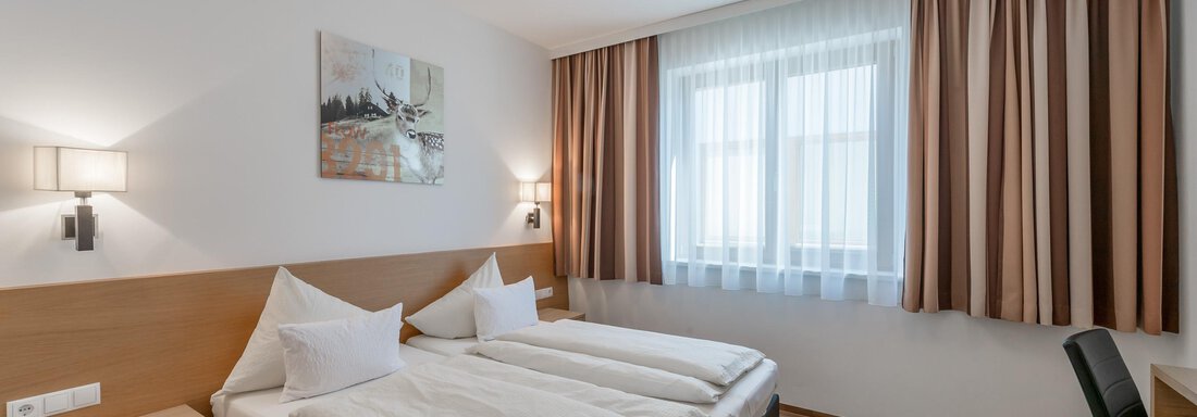 A modern bedroom with two beds, wooden flooring, and a large window front. The walls are painted light and decorated with curtains in neutral colors. - Apart Hotel Garni Wieser - Sölden
