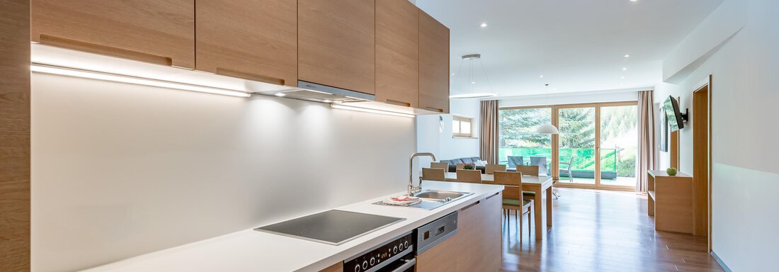 A modern kitchen with wood cabinets and a bright workspace. Large windows offer a view of a garden. - Apart Hotel Garni Wieser - Sölden