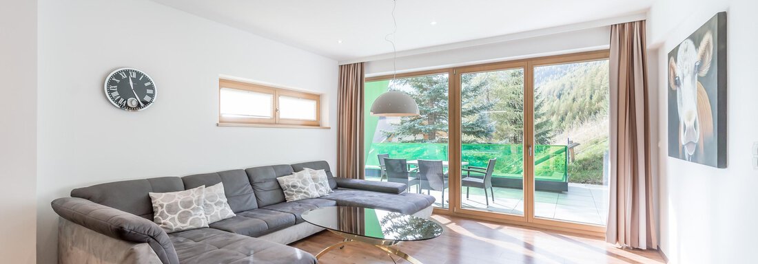 A modern living room with a gray sofa and a glass table. Large windows let in plenty of light and offer a view of the terrace. - Apart Hotel Garni Wieser - Sölden