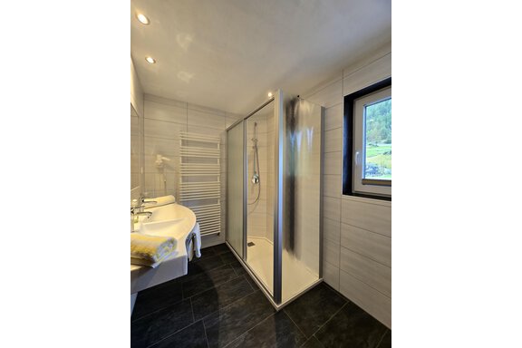A modern bathroom with a glass shower and a large window. The walls are tiled in light colors and some towels are visible. - Apart Tyrolis - Sölden