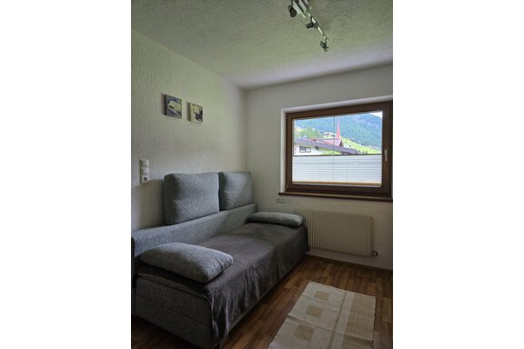 A cozy living room with a gray sofa and a window overlooking a green mountain landscape. Two small pictures hang on the wall. - Apart Tyrolis - Sölden