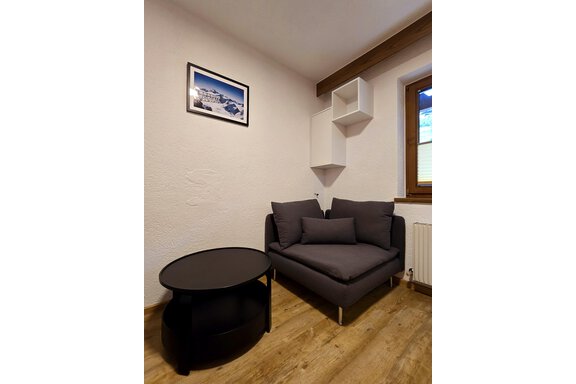 A cozy seating area with a gray sofa and a round table. A picture of a landscape hangs on the wall, and a window lets in natural light. - Apart Tyrolis - Sölden