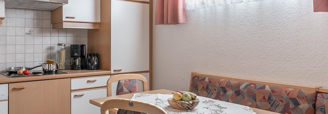 A modern kitchen with a dining table and a comfortable seating area. Fresh fruits are placed on the table and the windows are adorned with bright curtains. - Apart Wiesengrund - Sölden