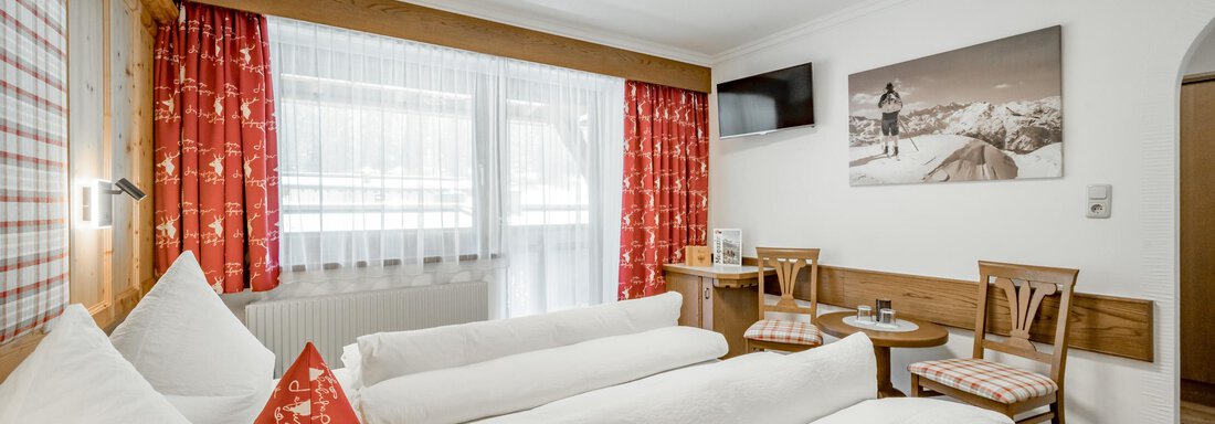 A cozy hotel room with two beds and light wood decor. The windows are adorned with red curtains and a TV is mounted on the wall. - Apart Wiesengrund - Sölden