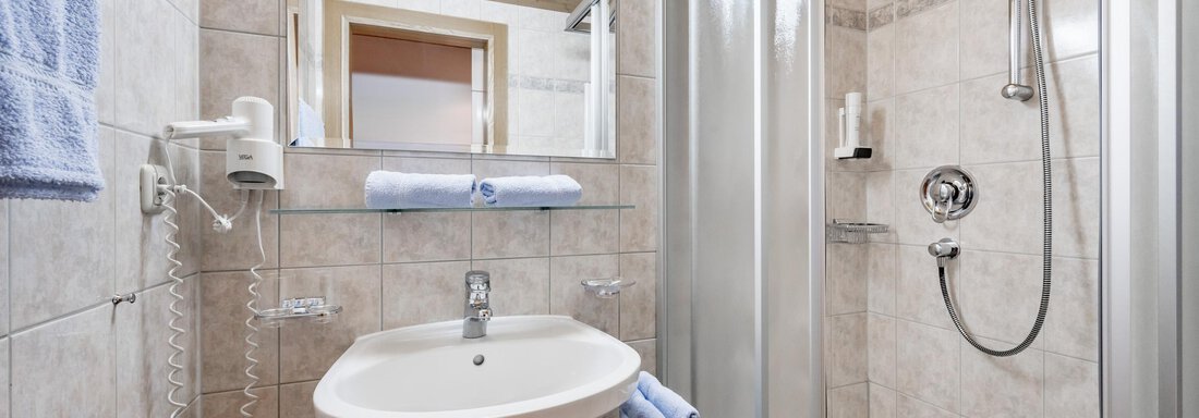 A modern bathroom with a shower, sink, and a large mirror. The walls are tiled in light colors, and there are fresh towels in blue. - Apart Wiesengrund - Sölden