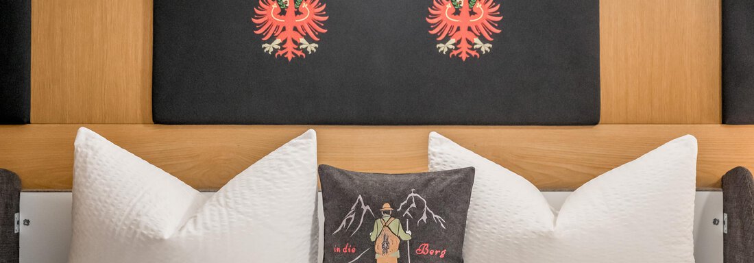 A cozy bedroom with white pillows and a black wall decorated with red emblems. On the bed lies a decorative pillow with a mountain motif. - Apart Wiesengrund - Sölden