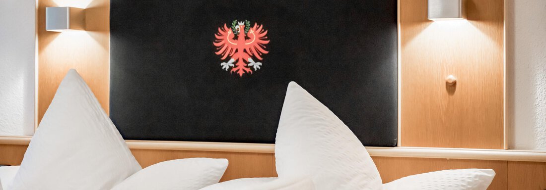 A cozy bedroom with a large headboard and decorative pillows. A stylized coat of arms can be seen on the wall. - Apart Wiesengrund - Sölden
