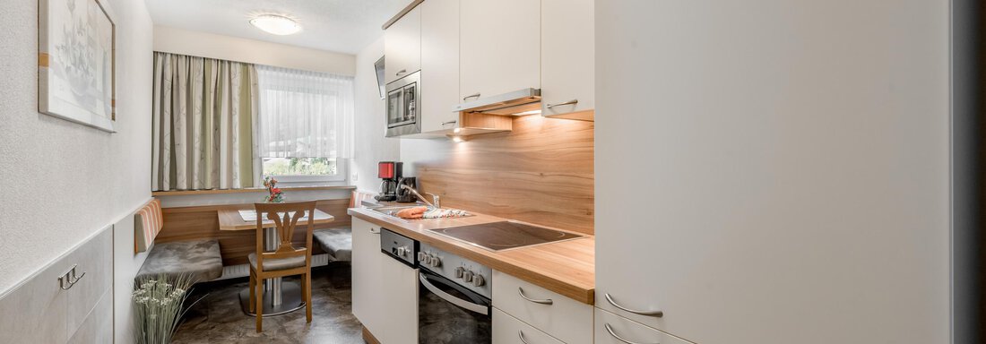 A modern kitchen with white cabinets and wooden details. There is a dining area with a table and benches, as well as large windows that let in natural light. - Apart Wiesengrund - Sölden