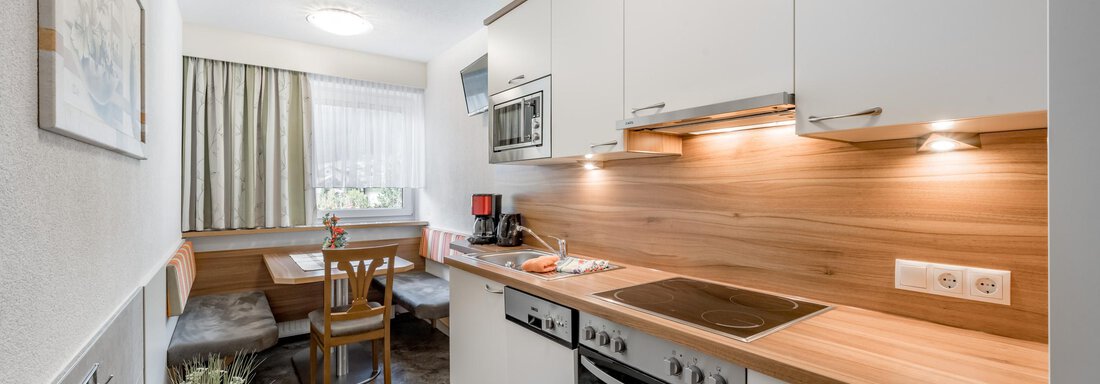 Kitchen A modern kitchen with white cabinets and wooden details. In the background, there is a small dining area with a table and chairs. - Apart Wiesengrund - Sölden