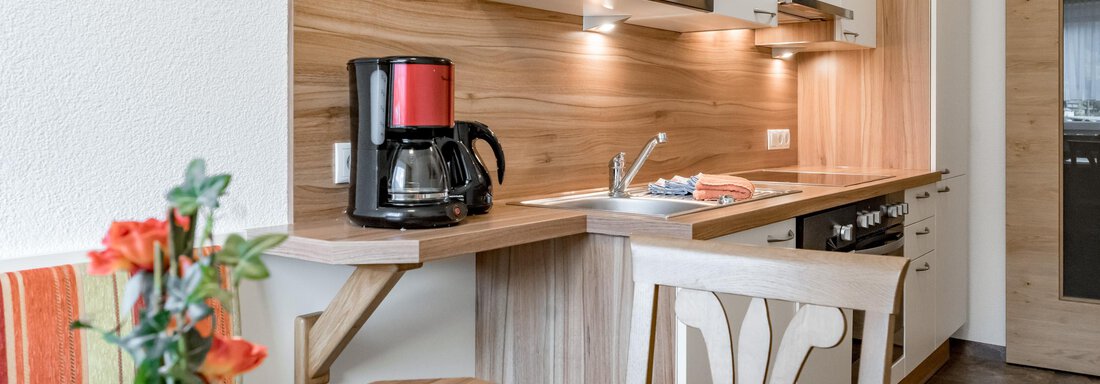 A modern kitchen with wood paneling, a kettle, and a microwave. On the table, there is a floral decoration and a freshly set dining table. - Apart Wiesengrund - Sölden