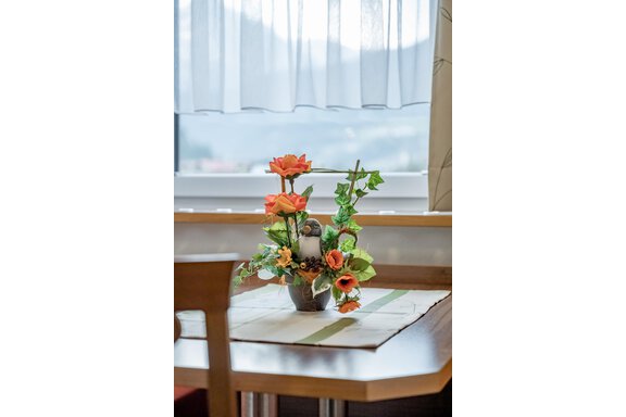 A beautiful table with a vase full of artificial flowers and a small bird. In the background, there are large windows with curtains and a picturesque view. - Apart Wiesengrund - Sölden