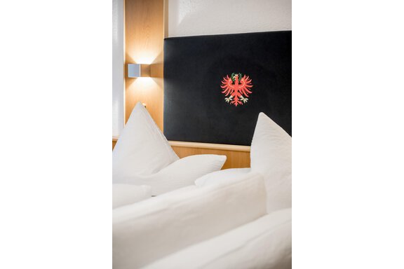 A cozy bed with large, white pillows.  
On the wall, there is a red coat of arms with a double eagle. - Apart Wiesengrund - Sölden