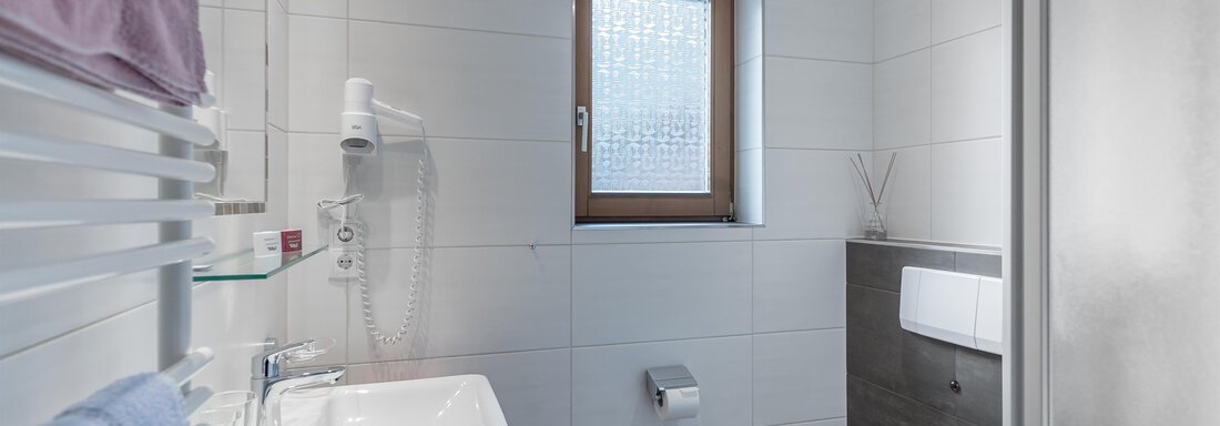 A modern bathroom with white tiles and a window. There is a sink, a toilet, and towels in soft colors. - Apart Wiesengrund - Sölden