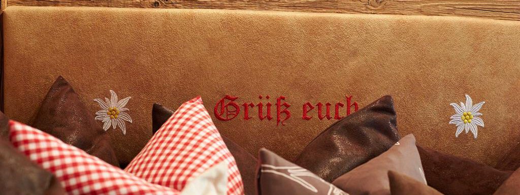 Ein gemütliches Kopfteil mit Kissen in verschiedenen Farben und Mustern. Darauf steht in roter Schrift "Grüß euch" mit einer dekorativen Blume. - Alpen Diamond Top 100 | 300 | 700 - Sölden