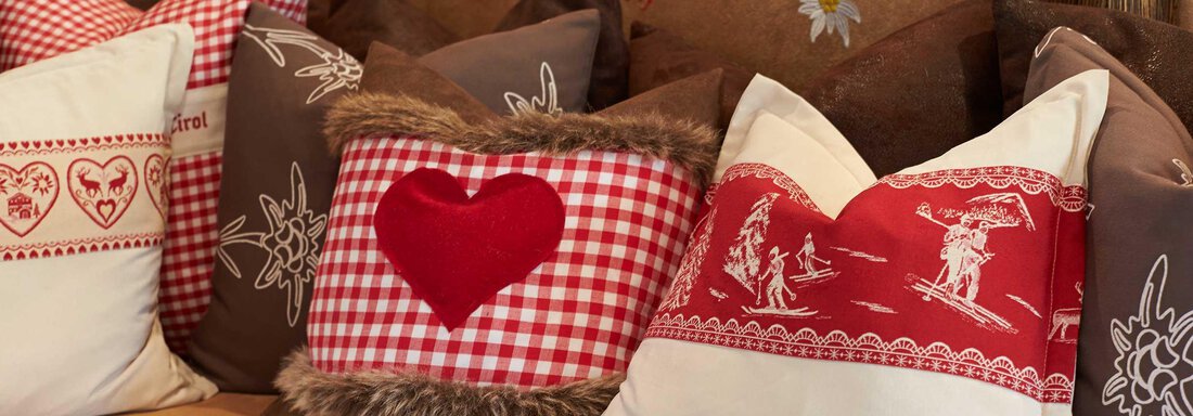 Eine gemütliche Ansammlung von Kissen in verschiedenen Designs. Die Kissen sind rot, braun und beige mit Herzmotiven und traditionellen Mustern. - Alpen Diamond Top 100 | 300 | 700 - Sölden