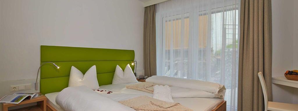 Ein modernes Hotelzimmer mit einem Doppelbett und einer grünen Kopfstütze. Große Fenster sorgen für viel Tageslicht und eine einladende Atmosphäre. - Alpen Diamond Top 100 | 300 | 700 - Sölden