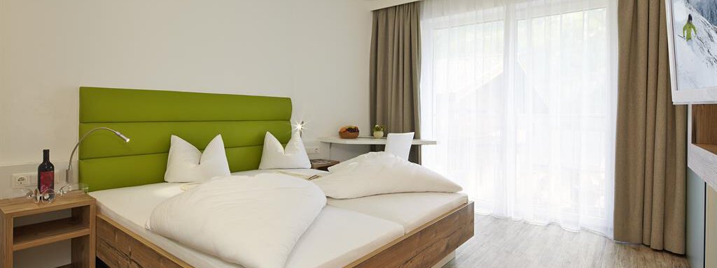 Ein modernes Schlafzimmer mit einem Doppelbett und grüner Wandverkleidung. Große Fenster sorgen für viel Licht und eine einladende Atmosphäre. - Alpen Diamond Top 100 | 300 | 700 - Sölden