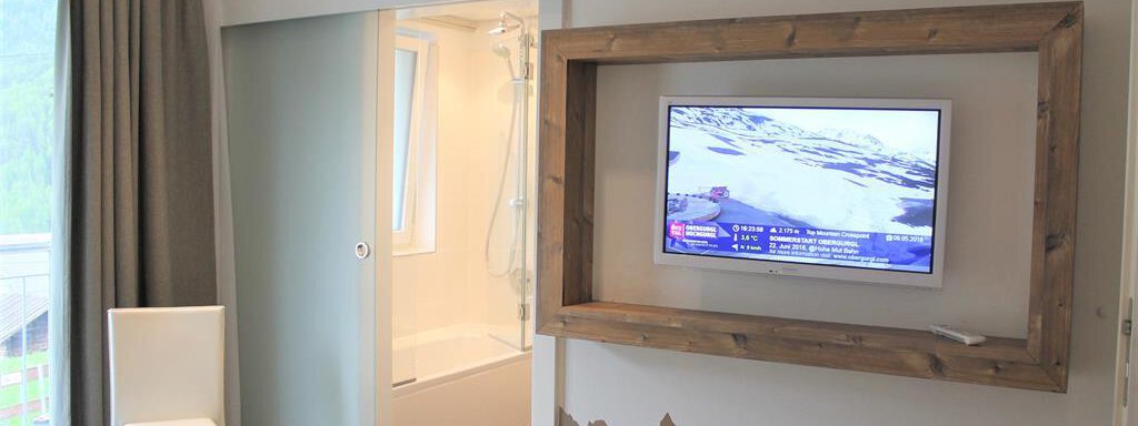 Ein modernes Zimmer mit einem Fernseher in einem Holzrahmen. Im Hintergrund sind ein Badezimmer und ein Fenster mit Vorhängen sichtbar. - Alpen Diamond Top 100 | 300 | 700 - Sölden
