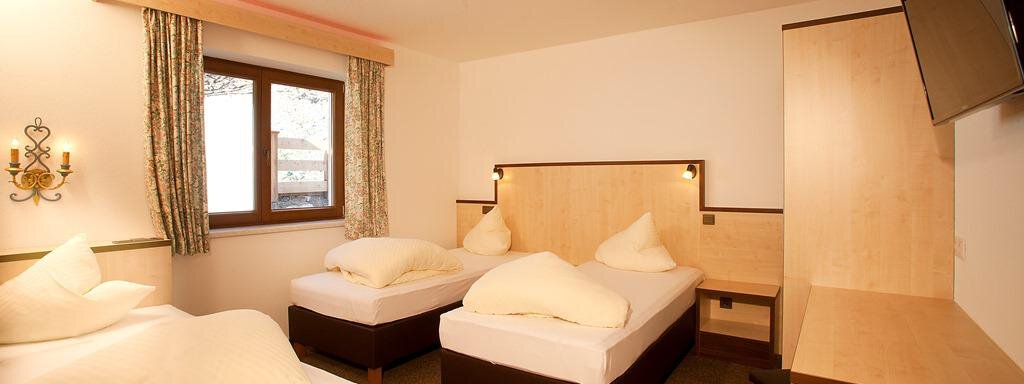 A cozy room with three beds and bright wooden furniture. The room features a window with curtains and a television. - Appartement Alpin - Sölden