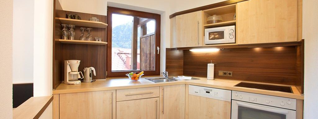 A modern kitchen with light wood cabinets and a window that looks out to the mountains. It is well-equipped with appliances such as a refrigerator, a stove, and a microwave. - Appartement Alpin - Sölden