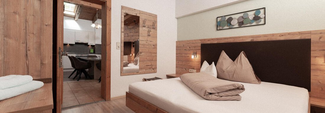A modern bedroom with wood-paneled walls and a large bed. The room is bright and cozy, with a view into an adjacent kitchen. - Appartement Gehörde Sölden - Sölden