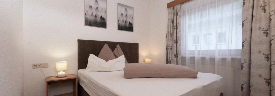 A cozy bedroom with a large bed and wooden ceiling. Two pictures hang on the wall, and the room has light curtains and warm light. - Appartement Gehörde Sölden - Sölden