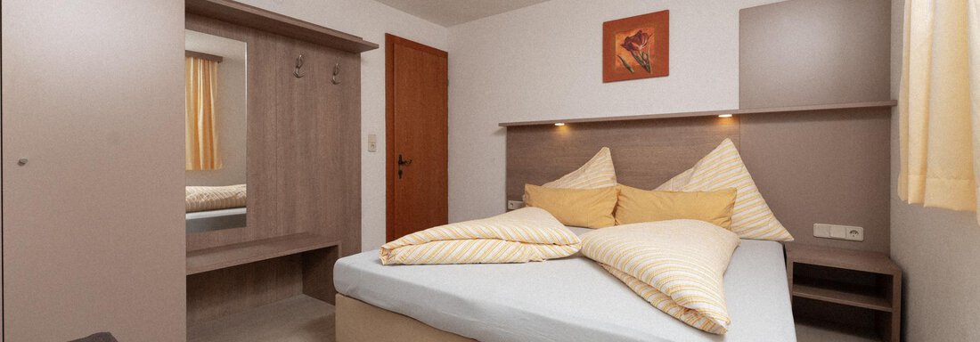 A modern bedroom with a double bed, two pillows, and a simple design. The walls are bright, and there is furniture like a wardrobe and a mirror. - Appartement Gehörde Sölden - Sölden