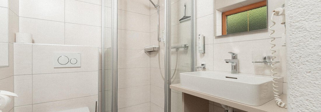 A modern bathroom with a shower, a sink, and a toilet. The walls are tiled in a light color and the furnishings are functional. - Appartement Gehörde Sölden - Sölden