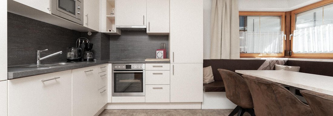 A modern kitchen with white cabinets and a dark countertop. There is a dining area with a cozy seating nook and a large table. - Appartement Gehörde Sölden - Sölden