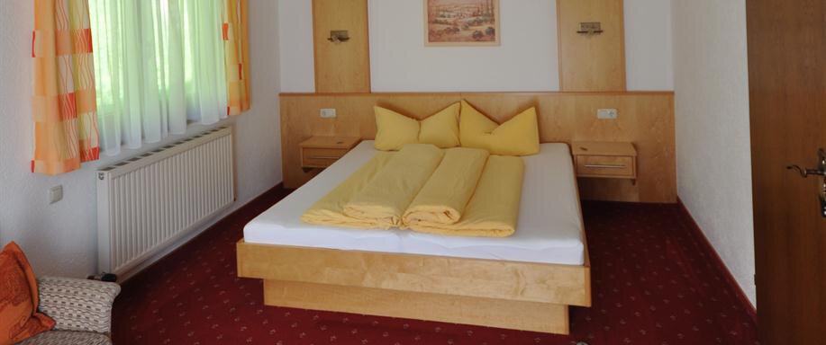 A cozy hotel room with a large double bed and yellow cushions. The room has a red carpet and a window with curtains. - Appartement Harmonie - Sölden