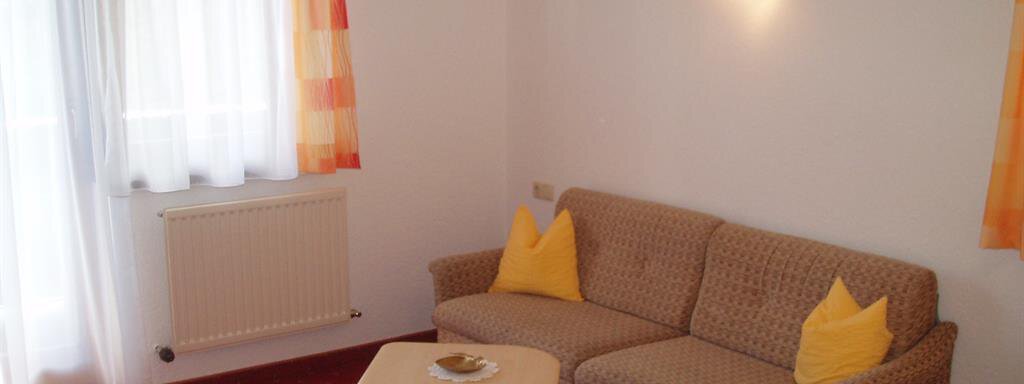 A cozy living room with a beige sofa and yellow pillows. Bright curtains let in a lot of light and the floor is covered with a red carpet. - Appartement Harmonie - Sölden