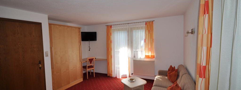 A cozy hotel room with a sofa, a table, and a television. The walls are light, and the curtains have an orange hue. - Appartement Harmonie - Sölden