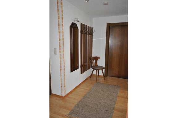 A simple hallway with a wooden chair, a coat rack, and a mirror. The floor is covered with a carpet and the walls are painted in light colors. - Appartement Harmonie - Sölden